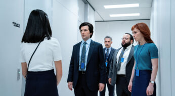 Auf dem Bild aus Severance Staffel 2 stehen von links nach rechts Miss Huang, gespielt von Sarah Bock, Mark Scout, gespielt von Adam Scott, Irving Bailiff, gespielt von John Turturro, Dylan George, gespielt von Zach Cherry, und Helly Riggs, gespielt von Britt Lower, in einem sterilen Büroflur. Alle tragen Bürokleidung mit blauen Namensschildern. Miss Huang steht mit dem Rücken zur Kamera, während die anderen ernsthaft und konzentriert wirken. Die weiße Umgebung und das Wort „Break Room“ an der Wand betonen die kontrollierte und angespannte Stimmung.