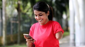 Junge Frau schaut lächelnd auf ein Smartphone, das sie in der rechten Hand hält.