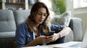 Junge Frau sitzt auf dem Boden vor ihrem Sofa und schaut lächelnd auf ihr Android-Handy.