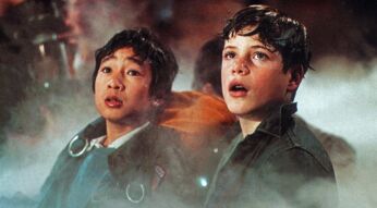Auf dem Bild in dem Artikel zur Die Goonies-Fortsetzung sind Ke Huy Quan als Data und Sean Astin als Mikey in einer spannenden Szene zu sehen. Beide blicken überrascht und ehrfürchtig in die Ferne, während dichter Nebel die Umgebung umhüllt. Data trägt eine Jacke mit Riemen, während Mikey in einem dunklen Hemd zu sehen ist. Die Szene strahlt Abenteuer und Entdeckung aus.