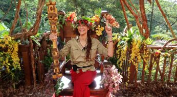 Auf dem Bild zum Dschungelcamp 2025 ist Lilly Becker als frisch gekrönte Dschungelkönigin zu sehen. Sie sitzt auf einem hölzernen Thron, trägt eine bunte Blumenkrone und hält das Siegerzepter in der Hand. Mit einem strahlenden Lächeln und erhobenem Finger feiert sie ihren Sieg. Der Thron ist mit bunten Blumen und Pflanzen geschmückt, im Hintergrund ist dichter, grüner Dschungel zu sehen. Die Stimmung ist feierlich und triumphierend.