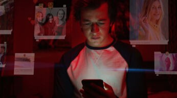 Das Bild zeigt eine Szene aus einem Film über Medienkonsum. Ein junger Mann sitzt in einem dunklen Raum und blickt konzentriert auf sein Smartphone, das sein Gesicht beleuchtet. Rote Lichteffekte verstärken die intensive Stimmung. Um ihn herum schweben digitale Social-Media-Posts mit Bildern, Likes und Kommentaren, die die allgegenwärtige Präsenz sozialer Netzwerke symbolisieren. Die Szene vermittelt eine kritische Auseinandersetzung mit digitaler Abhängigkeit und Online-Identität.