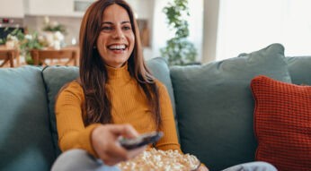 Eine junge Frau sitzt auf dem Sofa und hält eine Fernbedienung in der Hand.