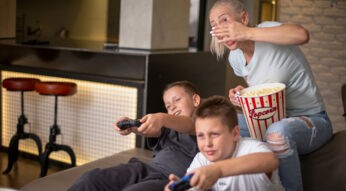 Zwei Jungen sitzen auf einem Sofa und spielen Mario Kart 9 auf der Nintendo Switch 2. Beide halten Controller und wirken konzentriert, während eine Frau hinter ihnen sitzt, Popcorn aus einer großen, rot-weiß gestreiften Schüssel hält und lachend mit der Hand ihr Gesicht teilweise bedeckt. Die Stimmung ist lebendig und entspannt, passend zu einer fröhlichen Spielrunde.