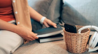 Eine Frau legt ihr Smartphone in einen kleinen geflochtenen Korb, der neben weißen Kopfhörern auf einem Sofa steht. Stattdessen hält sie ein Buch in der Hand, bereit zu lesen. Die Szene symbolisiert das Konzept "Phone Life Balance" – den bewussten Verzicht auf digitale Ablenkungen zugunsten analoger Aktivitäten und Entspannung.