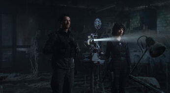 Auf dem Bild zu The Gorge Ende erklärt sind Anya Taylor-Joy als Drasa und Miles Teller als Levi in einer düsteren, verfallenen Umgebung zu sehen. Die beiden tragen taktische schwarze Kleidung und stehen vor einem alten Filmprojektor, der einen Lichtstrahl durch den staubigen Raum wirft. Levi blickt ernst in die Ferne, während Drasa konzentriert auf das projizierte Bild schaut. Die Szenerie wirkt geheimnisvoll und angespannt, verstärkt durch die spärliche Beleuchtung und die unheimliche Atmosphäre des verlassenen Ortes.