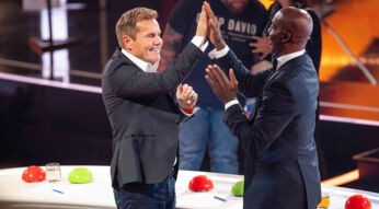Auf dem Bild in dem Artikel zu den Supertalent-Gewinner:innen sind zwei Juroren zu sehen. Der linke Juror, ein Mann mit kurzen blonden Haaren, trägt ein schwarzes Sakko und ein weißes Hemd, während er lächelnd die Hand hebt. Der rechte Juror, ein Mann mit Glatze im dunklen Anzug, erwidert das High-Five mit begeisterter Mimik. Im Hintergrund ist die Bühne mit leuchtenden Farben und Zuschauer:innen zu erkennen.