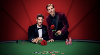 Auf dem Bild zu Die Bachelors 2025 stehen zwei stilvoll gekleidete Männer an einem Pokertisch mit grüner Filzfläche. Beide tragen elegante Samt-Sakkos, der linke in Dunkelblau mit Fliege, der rechte in Weinrot. Beide halten jeweils eine rote Rose in der Hand. Auf dem Tisch liegen Pokerchips und Würfel, der Hintergrund ist rot inszeniert.
