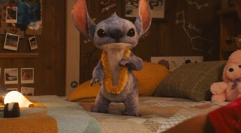 Auf dem Bild aus der Lilo und Stitch Live-Action-Adaption 2025 ist das blaue Alien Stitch in einer realistischen CGI-Version zu sehen. Er steht auf einem Bett, trägt eine gelbe Blumenlei und blickt direkt in die Kamera.