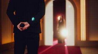 Auf dem Bild in dem Artikel zu Love Is Blind: Germany Staffel 2 steht ein Mann im schwarzen Anzug mit dem Rücken zur Kamera auf einem roten Teppich und hält eine kleine schwarze Schachtel hinter dem Rücken. Im Hintergrund erscheint eine Frau im Abendkleid.