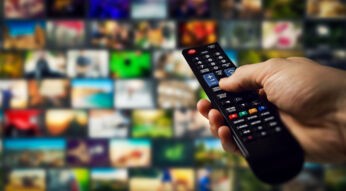 Eine Hand hält eine schwarze Fernbedienung und navigiert durch eine Auswahl an Streamingdiensten in Deutschland. Im Hintergrund sind zahlreiche unscharfe Bildschirmvorschauen von Filmen und Serien zu sehen, die eine große Vielfalt an Inhalten darstellen. Die Szene vermittelt das moderne Fernseherlebnis mit On-Demand-Streaming.