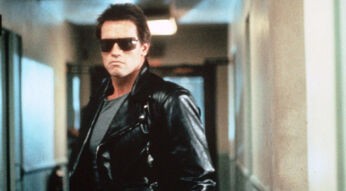 Auf dem Bild aus Terminator ist Arnold Schwarzenegger in seiner ikonischen Rolle als der Terminator zu sehen. Er trägt eine schwarze Lederjacke, eine dunkle Sonnenbrille und ein graues Shirt, während er mit ernstem Blick durch einen Flur geht. Die düstere Beleuchtung und sein ausdrucksloses Gesicht verstärken die bedrohliche Atmosphäre der Szene.