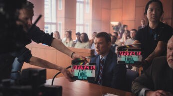 Auf diesem Bild zur Serie Nur für Dein Leben ist Sam Worthington als David Burroughs in einer Gerichtsszene zu sehen. Er sitzt ernst und fokussiert am Tisch, umgeben von Filmcrew-Mitgliedern mit Klappe, was auf eine laufende Produktion hinweist. Die Atmosphäre ist angespannt, mit Statisten im Hintergrund, die ein authentisches Gerichtssetting verstärken.