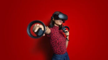 Eine junge Frau mit Virtual-Reality-Brille und zwei Controllern streckt den rechten Arm energisch nach vorne. Sie trägt ein rotes Oberteil mit Leopardenmuster vor knallrotem Hintergrund. Der Ausdruck wirkt fokussiert und spielerisch. Das Bild veranschaulicht auf moderne Weise die zentrale Frage: Was ist Virtual Reality?