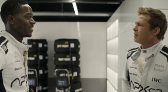 Bild aus F1: Zwei Rennfahrer in weißer Teamkleidung stehen in der Boxengasse und unterhalten sich lachend vor einem Stapel Rennreifen.