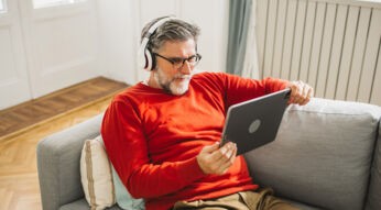 Ein älterer Mann mit grauem Haar und Bart sitzt entspannt auf einem hellgrauen Sofa und schaut mit Kopfhörern ein Video auf einem Tablet. Er trägt einen roten Pullover und wirkt konzentriert und zufrieden. Dieses Bild symbolisiert bequemes Streaming von zuhause und passt thematisch zur Darstellung der ARD Plus Kosten.