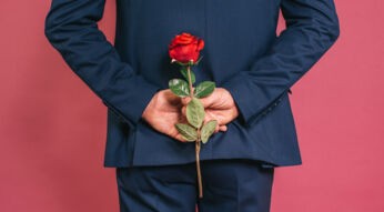 Auf dem Bild in dem Artikel zu den Bachelor-Formaten ist ein Mann in einem dunkelblauen Anzug von hinten zu sehen. Er hält eine rote Rose versteckt hinter seinem Rücken, während er vor einem pinkfarbenen Hintergrund steht.