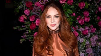 Auf dem Eventfoto ist Lindsay Lohan mit offenen, gewellten roten Haaren und einem schimmernden, bronzefarbenen Kleid zu sehen. Sie steht vor einer üppigen Blumenwand aus violetten und rosafarbenen Rosen und schaut direkt in die Kamera. Das Bild wird im Zusammenhang mit der möglicherweise geplanten Serie Count My Lies verwendet
