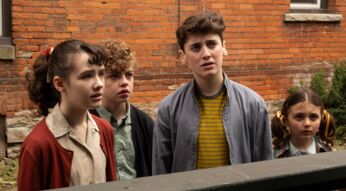 Welcome to Derry zeigt in dieser Szene vier Kinder, die mit ernsten und teils verunsicherten Blicken in dieselbe Richtung schauen. Die Gruppe steht vor einer Backsteinwand, ihre Kleidung wirkt retro und deutet auf eine frühere Zeitperiode hin.