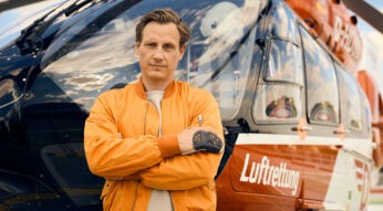 Auf dem Bild zu den ZDF-Serien Empfehlungen ist Schauspieler Patrick Kalupa als Dr. Moritz Neiss zu sehen. Er steht mit verschränkten Armen und ernstem Blick vor einem Rettungshubschrauber mit der Aufschrift „Luftrettung“. Kalupa trägt eine orangefarbene Fliegerjacke, was seine Rolle als Notarzt unterstreicht. Der Himmel spiegelt sich dramatisch in der Cockpit-Scheibe.