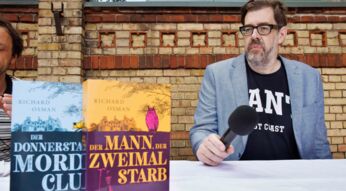 Der Bestsellerautor Richard Osman sitzt mit seinen "Der Donnerstagsmordclub"-Büchern an einem Tisch.