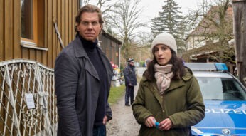 Auf dem Bild zu Der Alte Staffel 50 stehen Caspar Bergmann (Thomas Heinze) und Annabell Lorenz (Stephanie Stumph) vor einem Schuppen an einem Tatort. Beide wirken aufmerksam und nachdenklich, während sie sich austauschen. Im Hintergrund steht ein Polizeiwagen sowie ein Beamter, was die angespannte, kriminalistische Atmosphäre der Szene unterstreicht.