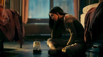 In dieser melancholischen Szene zur John Wick Ballerina Kritik sitzt Ana de Armas als Eve gedankenverloren auf dem Boden eines Schlafsaals zwischen zwei Betten. Sie trägt gemütliche Kleidung und blickt traurig auf eine Spieluhr mit einer tanzenden Figur unter einer Glaskuppel. Das warme Licht unterstreicht die emotionale Tiefe des Moments.