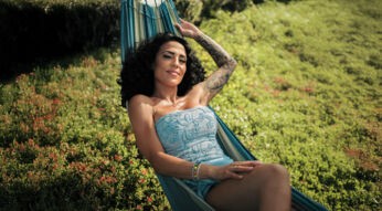 Auf dem Bild zu Make Love Fake Love Staffel 4 liegt Elena Miras entspannt in einer Hängematte. Sie trägt ein hellblaues, schulterfreies Outfit mit Blumenstickerei, ihr lockiges Haar fällt über die Schultern. Der Hintergrund zeigt eine grüne Wiese mit kleinen roten Blüten.