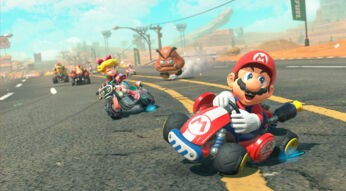 Auf diesem Bild zu Mario Kart World Tipps driftet Mario fröhlich mit seinem roten Kart durch eine Wüstenlandschaft. Hinter ihm fährt Peach auf einem Motorrad und zwinkert in die Kamera, während weitere Fahrer und ein Goomba die Szene beleben. Die Grafik ist detailreich und dynamisch, was die rasante Rennatmosphäre perfekt einfängt.