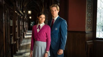 Auf dem Bild zur "Maxton Hall" Staffel 3 sind Harriet Herbig-Matten als Ruby Bell und Damian Hardung als James Beaufort in ihren Schuluniformen zu sehen. Ruby trägt eine pinke Strickjacke und einen grauen Faltenrock, James einen blauen Blazer mit Wappen. Beide stehen in einem historischen Schulflur mit dunklem Holz und wirken ernst und fokussiert.