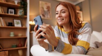 Rothaarige Frau liegt auf dem Sofa und schaut auf ihr iPhone