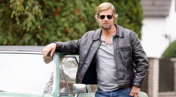 Auf dem Bild in dem Artikel zur Besetzung von Der letzte Bulle steht ein Mann mit blonder Frisur und Bart lässig an einem grünen Oldtimer. Er trägt eine schwarze Lederjacke, ein graues Shirt und eine Sonnenbrille.