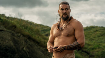 In dieser Szene aus der Chief of War-Serie ist Ka'iana, gespielt von Jason Momoa, zu sehen. Er steht oberkörperfrei in einer natürlichen, grünen Landschaft, trägt eine traditionelle Kette aus Zähnen und hat markante polynesische Tattoos am Arm und Bein. Der ernste Blick und die kraftvolle Körperhaltung verleihen der Szene aus der Chief of War-Serie eine entschlossene, kämpferische Stimmung.