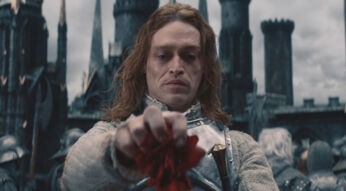 In einer Schlüsselszene aus Dracula - Die Auferstehung steht Dracula, gespielt von Caleb Landry Jones, in silberner Rüstung vor einer düsteren Burgkulisse. Mit ernstem Blick hält er eine rote Blume in der Hand, als Symbol für Verlust oder Erinnerung. Der Himmel ist grau, die Stimmung bedrohlich und melancholisch zugleich.