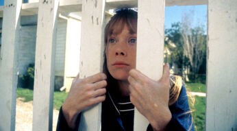 Carrie schaut durch einen Lattenzaun. Sie kniet und hält sich daran fest.