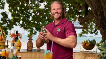 Das Bild zu Bachelor in Paradise 2025 zeigt Paul Janke, der an einer Außenbar steht. Er trägt ein bordeauxrotes Polohemd mit einem grünen Palmen-Logo und lächelt in die Kamera.