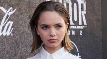 Auf dem Bild in dem Artikel zum Buffy Reboot ist Schauspielerin Ryan Kiera Armstrong zu sehen. Sie steht vor einer dunklen Fotowand, trägt ein weißes Hemd und blickt ernst in die Kamera.