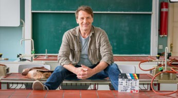 Im Bild zur Serie Der Lehrer bei RTL sitzt ein Mann auf einem Schultisch vor einer grünen Tafel. Neben ihm liegt eine Lederumhängetasche, vor ihm steht eine Filmklappe mit dem Serientitel.