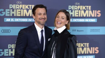Auf diesem Bild zur symbolischen Ankündigung von Der perfekte Urlaub sind Florian David Fitz und Jessica Schwarz bei der Premiere von Das perfekte Geheimnis zu sehen. Beide Schauspieler lächeln herzlich in die Kamera. Fitz trägt einen dunklen Anzug mit Krawatte, während Schwarz mit weißem Hemd und schwarzem Blazer auffällt. Die Stimmung wirkt gelöst und fröhlich.