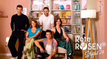 Auf dem Bild zur Rote Rosen Staffel 24 posieren fünf Schauspieler:innen vor einem hellen Bücherregal. Zwei Personen sitzen auf einem Sofa, drei stehen oder sitzen locker drumherum.