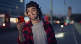 Im Bild zur Garantie bei Apple siehst du einen jungen Mann mit dunklen Locken und roter Handyhülle, der in der Abenddämmerung lächelnd auf sein iPhone schaut.