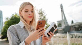 Frau steht in Großstadt in der Nähe einer Brücke und hält in einer Hand ein iPhone, in der anderen eine Powerbank