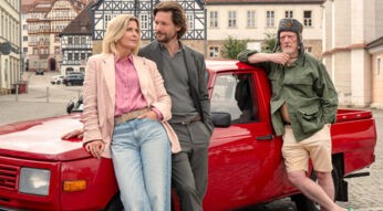 Auf dem Bild zu den neue Folgen von Nächste Ausfahrt Glück lehnt Katharina, gespielt von Valerie Niehaus, entspannt an einem roten Pickup. Neben ihr steht eine männliche Figur in grauer Kleidung. Rechts lehnt Willi, gespielt von Ernst Stötzner, lässig an der Ladefläche, mit grüner Jacke, Shorts und Fellmütze. Die Szene wirkt locker und freundlich, im Hintergrund sind Fachwerkhäuser einer deutschen Altstadt zu sehen.