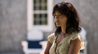 Auf dem Bild zur Serie Solange wir lügen Staffel 2 ist Emily Alyn Lind in ihrer Rolle als Cadence Sinclair Eastman zu sehen. Sie steht leicht seitlich im Bild, mit ernster Miene und nachdenklichem Blick. Ihr cremefarbenes Kleid mit Rüschen und das dunkle, leicht zerzauste Haar betonen den dramatischen Ton der Szene. Der Hintergrund zeigt unscharfe Gartenmöbel vor einer Hausfassade.