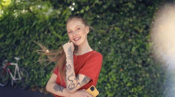 Android Datenverbrauch: Junge Frau in rotem T-Shirt steht mit ihrem Smartphone vor einer Hecke.