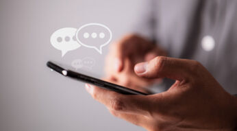Eine Person hält ein Smartphone in den Händen, Sprachblasen symbolisieren Kommunikation via Messenger.