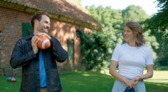 Auf dem Bild zur Besetzung von Rote Rosen in Staffel 24 sind Daniel, gespielt von Daniel Fritz, und Lou, dargestellt von Eva-Maria Grein von Friedl, in einer sommerlichen Szene im Grünen zu sehen. Beide blicken sich lächelnd an, während Daniel einen Football hält. Die Stimmung wirkt leicht und verspielt, mit einem ländlichen Gebäude im Hintergrund.