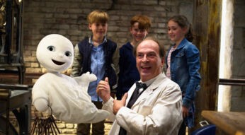 Auf dem Bild aus dem Film Das kleine Gespenst, einem Klassiker unter den Halloween-Filmen für die ganze Familie, ist das fröhlich dreinblickende Gespenst mit einem großen Schlüsselbund in der Luft schwebend zu sehen. Daneben steht ein lachender Mann in weißem Kittel, umgeben von drei neugierigen Kindern vor einer rustikalen Ziegelwand. Die Stimmung ist heiter und kindgerecht.