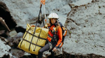 Katharina Strasser (Luise Bähr) hängt vor einer Felswand an einem Rettungstau. Ebenfalls am Tau ist ein gelber zusammengelegter Bergesack befestigt.