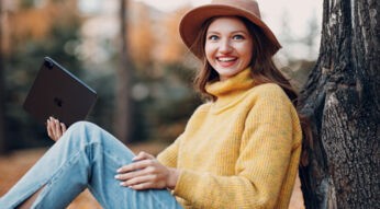 Ein junge Frau lehnt an einem Baum. Sie hält ein iPad in der Hand und lächelt in die Kamera.